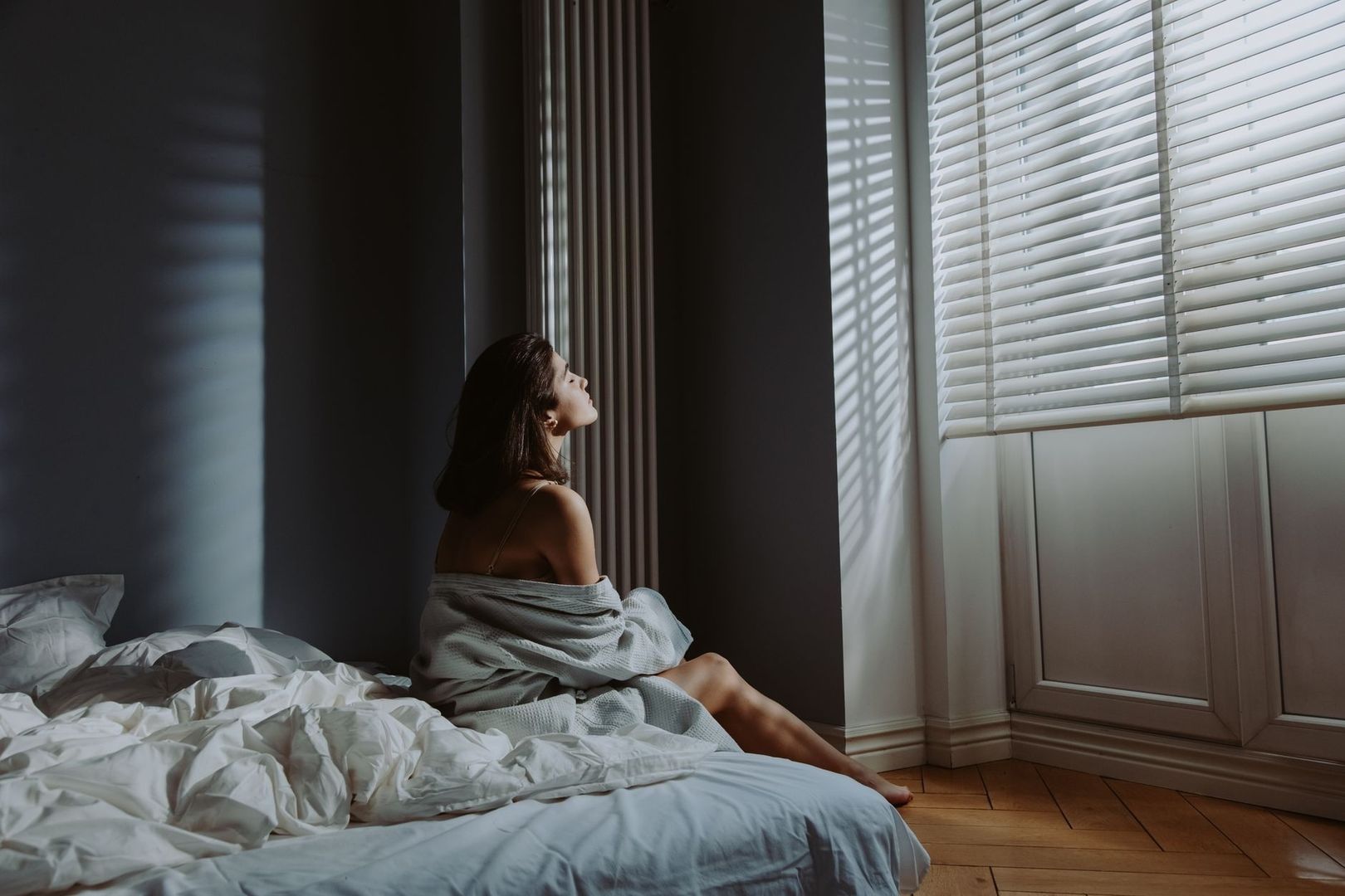 Kvinna sitter på sängen i morgonljus från fönstrets persienner